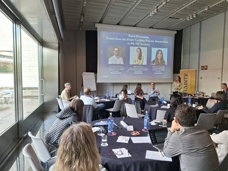 Panel discussion in a conference room with attendees seated at round tables and speakers on stage.